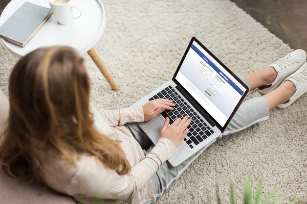 Vista de ángulo alto de la mujer en casa sentado en el suelo y el uso de ordenador portátil con el sitio web de facebook en la pantalla - foto de stock