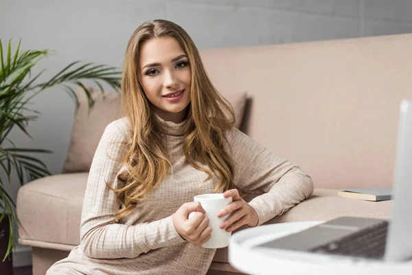 Schöne junge Frau, die zu Hause Kaffee trinkt, während sie mit Laptop arbeitet — Stockfoto