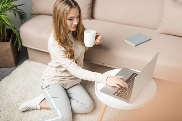 Jeune femme travaillant avec un ordinateur portable à la maison tout en étant assis sur le sol et en buvant du café — Photo de stock