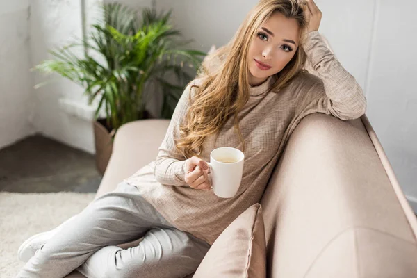 Attraente giovane donna con tazza di bevanda calda seduta sul divano — Foto stock