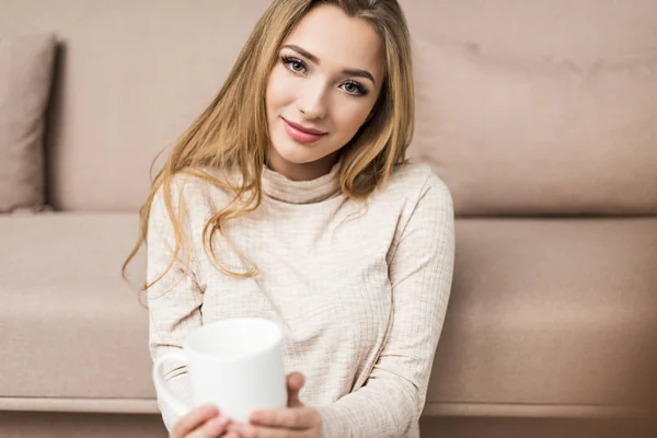 Schöne junge Frau im warmen Pullover mit einer Tasse Heißgetränk zu Hause — Stockfoto