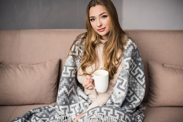 Beautiful young woman covered in warm plaid drinking coffee on couch — Stock Photo