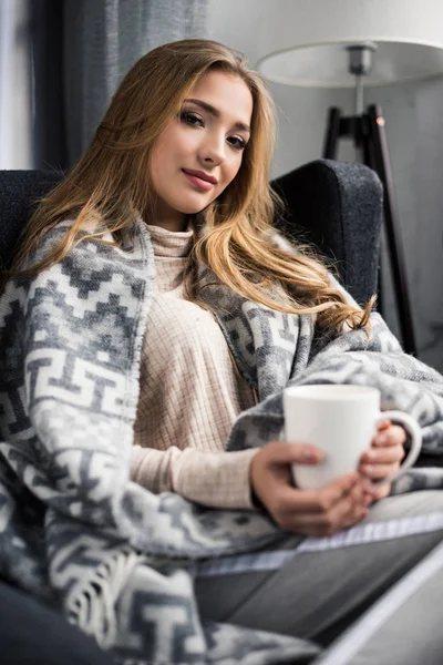 Happy young woman covered with warm plaid drinking coffee — Stock Photo