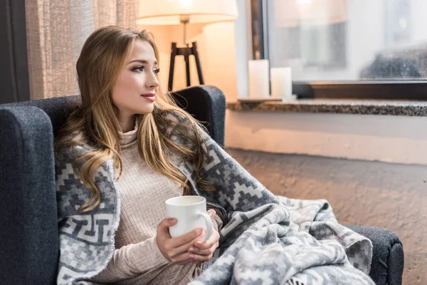 Réfléchie jeune femme en plaid chaud avec tasse de boisson chaude — Photo de stock