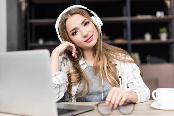 Glückliche junge Frau, die am Arbeitsplatz mit Kopfhörern Musik hört und in die Kamera schaut — Stockfoto