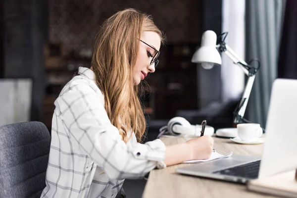 Konzentrierte junge Frau schreibt am Arbeitsplatz in Notizbuch — Stockfoto