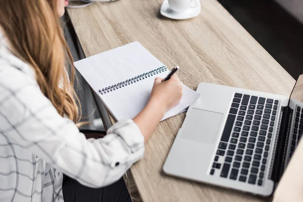 Tiro recortado de mulher jovem escrevendo em notebook no local de trabalho — Fotografia de Stock