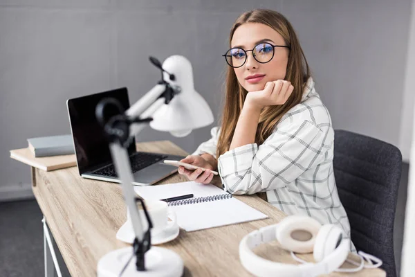 Young woman using smartphone at workplace and looking at camera — Stock Photo