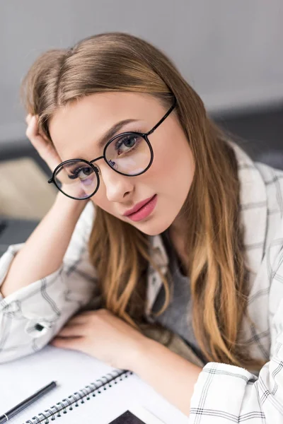 Hermosa chica cansada studert con portátil sentado en el lugar de trabajo y mirando a la cámara - foto de stock