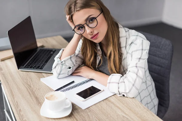 Exhausted young businesswoman sitting at workplace in office and looking at camera — Stock Photo