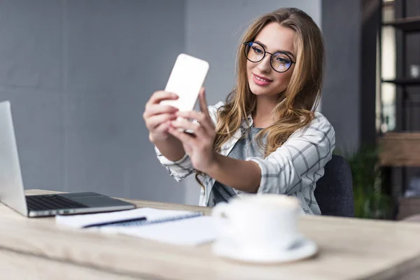 Smiling young businesswoman taking selfie at workplace — Stock Photo