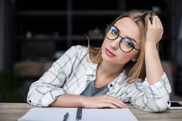 Junges müdes Mädchen mit Notizbuch sitzt am Arbeitsplatz und blickt in die Kamera — Stockfoto