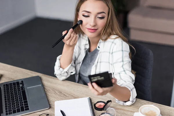 Attraktive junge Frau, die sich am Arbeitsplatz schminkt und in den Spiegel schaut — Stockfoto