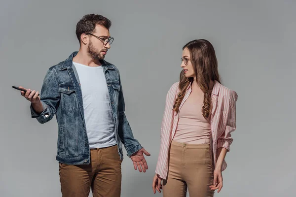 Stilvoller junger Mann mit Brille, Smartphone in der Hand und Freundin isoliert auf grau anschauend — Stockfoto