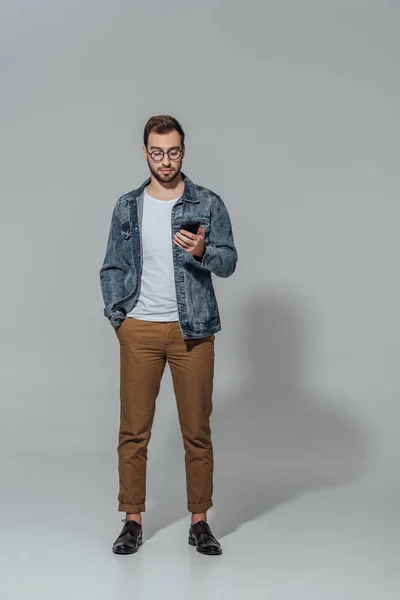 Vista completa de hombre joven elegante guapo en gafas con teléfono inteligente aislado en gris - foto de stock