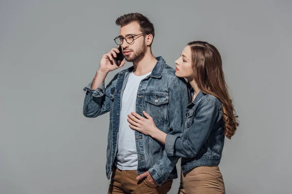 Jovem mulher tocando e olhando para o homem elegante em óculos falando no smartphone isolado no cinza — Fotografia de Stock