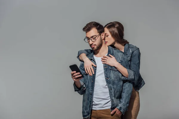 Junge Frau umarmt gutaussehenden Mann mit Brille und Smartphone isoliert auf grau — Stockfoto