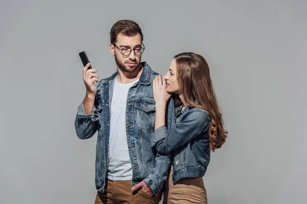 Junge Frau schaut gutaussehenden Mann mit Brille an und hält Smartphone isoliert auf grau — Stockfoto