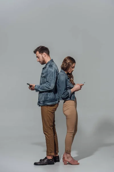 Full length side view of young couple in denim jackets standing back to back and using smartphones isolated on grey — Stock Photo