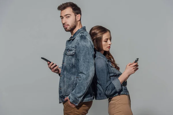 Молода пара в джинсових куртках, що стоять спиною до спини і використовують смартфони ізольовані на сірому — Stock Photo