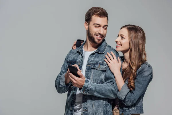 Красива молода пара в джинсових куртках використовує смартфони і посміхається один одному ізольовано на сірому — стокове фото