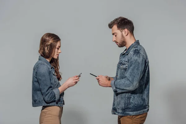 Vista lateral do jovem casal em jaquetas de ganga usando smartphones isolados em cinza — Fotografia de Stock