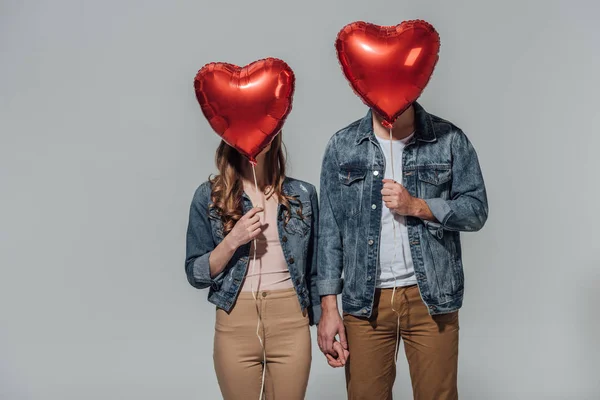 Jeune couple cachant des visages derrière des ballons rouges en forme de coeur isolés sur gris — Photo de stock