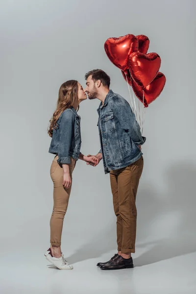 Vista lateral de pareja joven besándose mientras el hombre sostiene globos rojos en forma de corazón aislados en gris - foto de stock