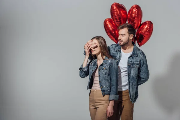 Junger Mann mit roten herzförmigen Luftballons und geschlossenen Augen vor glücklicher Freundin isoliert auf grau — Stockfoto