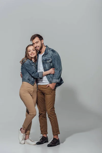 Вид в полный рост веселой молодой пары, обнимающейся и улыбающейся перед камерой на сером фоне — стоковое фото