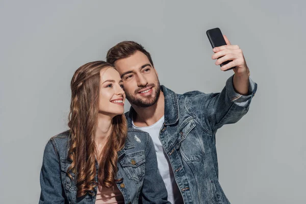 Happy young couple in denim jackets taking selfie with smartphone isolated on grey — Stock Photo