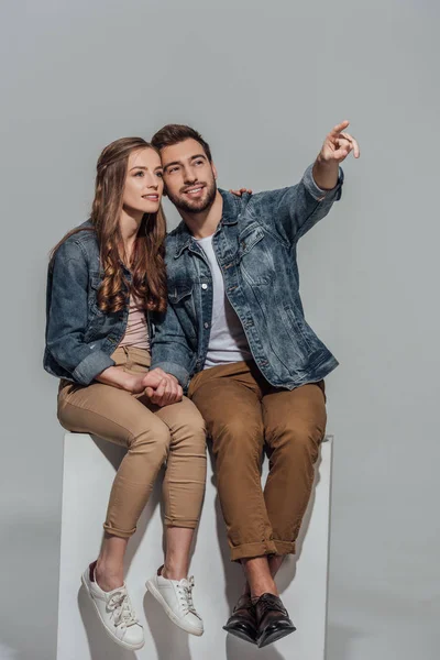 Belo casal jovem sorridente sentado junto e apontando para longe com o dedo isolado no cinza — Fotografia de Stock