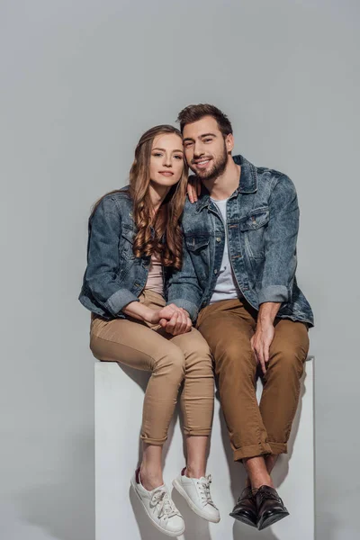 Hermosa pareja joven en chaquetas de mezclilla sentados juntos y sonriendo a la cámara aislada en gris - foto de stock