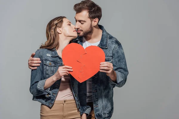 Young couple holding pieces of broken heart symbol together and kissing isolated on grey — Stock Photo
