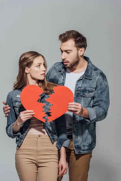 Chateado jovem casal segurando pedaços de símbolo de coração quebrado e olhando um para o outro isolado no cinza — Fotografia de Stock
