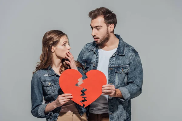 Upset young couple holding pieces of broken heart symbol isolated on grey — Stock Photo