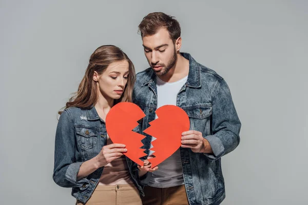 Joven pareja molesta sosteniendo partes del símbolo del corazón roto aisladas en gris - foto de stock