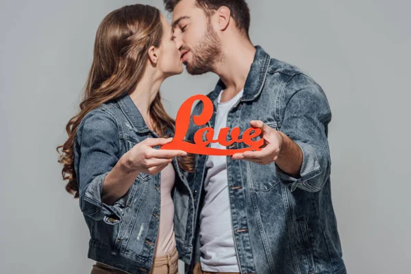 Nahaufnahme eines jungen Paares, das ein rotes Liebessymbol hält und sich isoliert auf grau küsst — Stockfoto