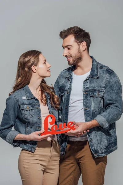 Feliz jovem casal segurando símbolo de amor vermelho e olhando um para o outro isolado no cinza — Fotografia de Stock