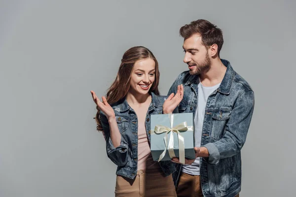 Bel giovane uomo che presenta scatola regalo alla fidanzata sorpresa isolata sul grigio — Foto stock