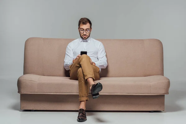 Hombre con estilo en gafas de vista sentado en el sofá y el uso de smarttphone aislado en gris - foto de stock