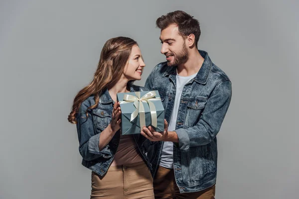 Красива щаслива молода пара тримає подарункову коробку і посміхається один одному ізольовано на сірому — стокове фото