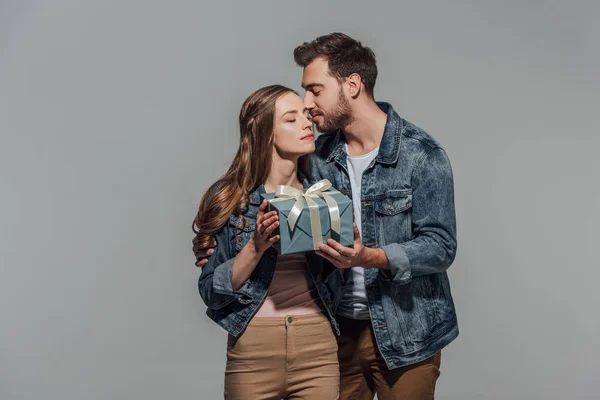 Schöner junger Mann küsst Freundin und präsentiert Geschenkbox isoliert auf grau — Stockfoto