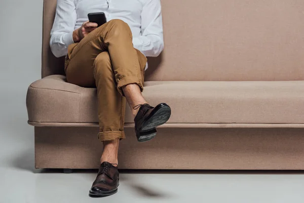 Sección baja de hombre joven usando teléfono inteligente mientras está sentado en el sofá aislado en gris - foto de stock