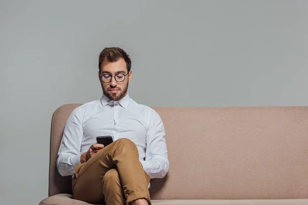 Jovem hansome em óculos usando smartphone enquanto sentado no sofá isolado em cinza — Fotografia de Stock