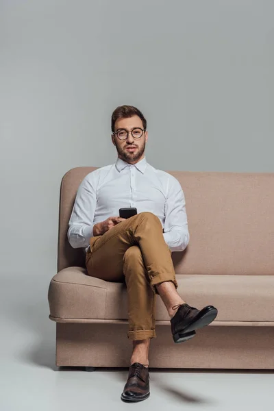 Junger Mann mit Brille hält Smartphone in der Hand und blickt in die Kamera, während er isoliert auf einem Sofa in grau sitzt — Stockfoto