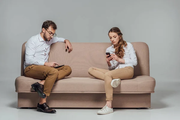 Joven pareja elegante sentado en el sofá y el uso de teléfonos inteligentes aislados en gris - foto de stock