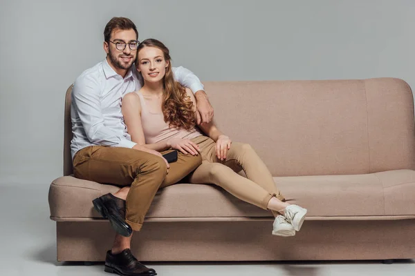 Feliz pareja joven y elegante con teléfono inteligente sonriendo a la cámara mientras se sientan juntos en el sofá aislado en gris - foto de stock