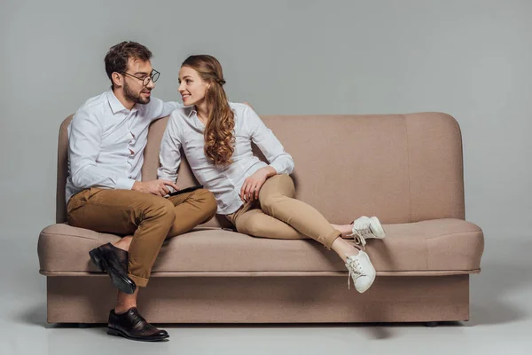 Красива усміхнена молода пара використовує смартфон разом, сидячи на дивані ізольовано на сірому — стокове фото