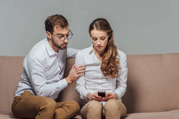 Grave jovem mulher usando smartphone enquanto homem bonito em óculos sentado no sofá isolado no cinza — Fotografia de Stock